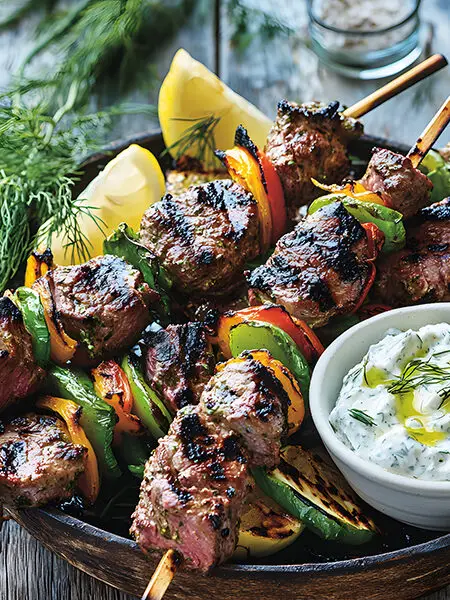 Juicy herb-marinated lamb souvlaki skewers with charred edges, grilled vegetables, and creamy tzatziki sauce on a rustic Mediterranean table