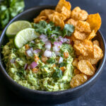 A bowl of creamy keto guacamole with fresh ingredients, served with crispy, golden chicharrónes for dipping.